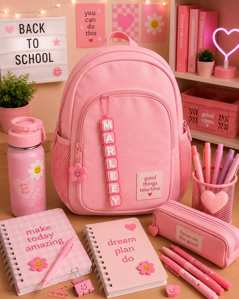 Op de afbeelding: Een roze rugzak met een gepersonaliseerd naamlabel met de tekst MARLEEY, een roze waterfles met een smiley, twee spiraalnotitieblokken met tekst, een roze etui en een roze pennenhouder van gaas met diverse roze pennen en stiften.