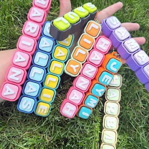 Op de afbeelding: Een verzameling kleurrijke, rechthoekige, blokvormige sleutelhangers met verhoogde witte letters. De getoonde namen zijn MARIANA, DALLAS, HALLIE, SAUL, LILY, HANNAH, JEYMI, SAMANTHA en HARBAE, elk bevestigd aan een kleine kralenketting.