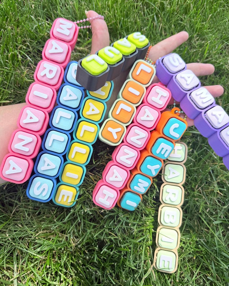 Op de afbeelding: Een verzameling kleurrijke, rechthoekige, blokvormige sleutelhangers met verhoogde witte letters. De getoonde namen zijn MARIANA, DALLAS, HALLIE, SAUL, LILY, HANNAH, JEYMI, SAMANTHA en HARBAE, elk bevestigd aan een kleine kralenketting.