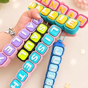 Op de afbeelding: Een collectie kleurrijke siliconen naamsleutelhangers met verhoogde witte letters op vierkante blokjes. De sleutelhangers tonen de namen HANNAH, HALLIE, HAILEY, SAUL, LISSETH en JUNIOR in diverse felle kleurcombinaties, waaronder roze, blauw, geel en zwart.