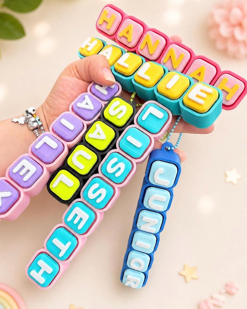 Op de afbeelding: Een collectie kleurrijke siliconen naamsleutelhangers met verhoogde witte letters op vierkante blokjes. De sleutelhangers tonen de namen HANNAH, HALLIE, HAILEY, SAUL, LISSETH en JUNIOR in diverse felle kleurcombinaties, waaronder roze, blauw, geel en zwart.