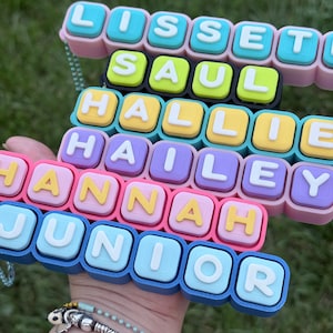 Op de afbeelding: Een veelkleurig naamplaatje-accessoire in blokstijl met gestapelde rijen individuele vierkante tegels. Elke tegel toont een enkele witte letter en spelt de namen LISSETTE, SAUL, HALLIE, HAILEY, HANNAH en JUNIOR in verschillende pastel- en felle kleuren.
