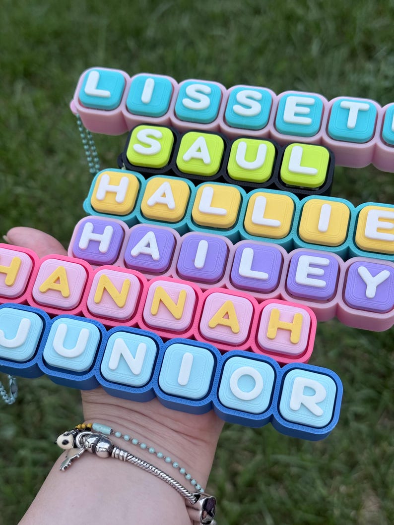 Op de afbeelding: Een veelkleurig naamplaatje-accessoire in blokstijl met gestapelde rijen individuele vierkante tegels. Elke tegel toont een enkele witte letter en spelt de namen LISSETTE, SAUL, HALLIE, HAILEY, HANNAH en JUNIOR in verschillende pastel- en felle kleuren.