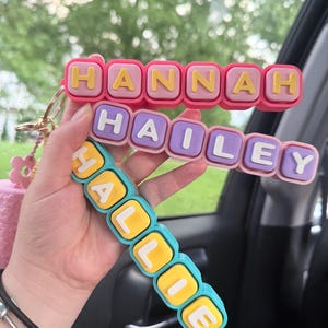 Op de afbeelding: Drie gestapelde siliconen sleutelhangers met blokletters die de namen HANNAH, HAILEY en HALLIE vormen. De bovenste sleutelhanger is roze met gele letters, de middelste is lichtpaars met witte letters en de onderste is blauwgroen met gele letters.