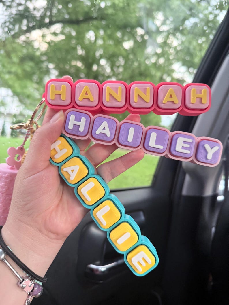 Op de afbeelding: Drie gestapelde siliconen sleutelhangers met blokletters die de namen HANNAH, HAILEY en HALLIE vormen. De bovenste sleutelhanger is roze met gele letters, de middelste is lichtpaars met witte letters en de onderste is blauwgroen met gele letters.