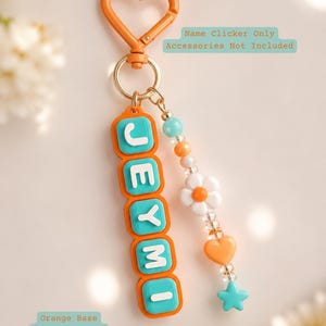 Op de afbeelding: Een oranje hartvormige sleutelhanger met een verticaal naamplaatje met de naam JEYMI in witte letters op turquoise dopjes en een oranje basis. Een kralensnoer met een witte bloem, een oranje hart en een turquoise ster hangt aan de ring.
