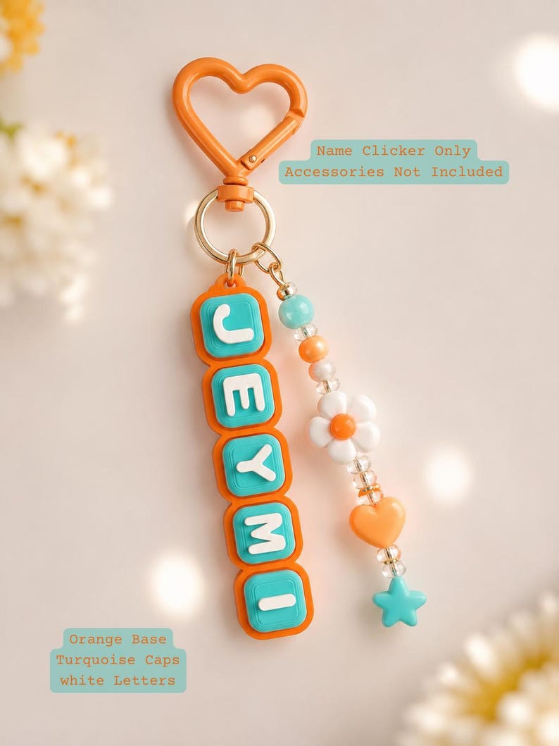 Op de afbeelding: Een oranje hartvormige sleutelhanger met een verticaal naamplaatje met de naam JEYMI in witte letters op turquoise dopjes en een oranje basis. Een kralensnoer met een witte bloem, een oranje hart en een turquoise ster hangt aan de ring.
