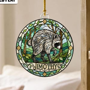 May include: A circular hanging ornament featuring a stained glass style illustration of a raccoon in a forest. The design includes green foliage, a blue sky, and the name JIMOTHY printed in black letters at the bottom. A gold cord is attached.