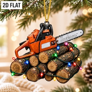 May include: A 2D flat hanging ornament featuring an orange chainsaw resting on a stack of brown logs. The chainsaw blade is printed with the name Jackson. A string of small, multi-colored Christmas lights is wrapped around the logs.