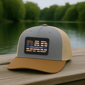 May include: A gray and tan baseball cap with a brown brim. The cap features a rectangular patch with the word "DAD" and a blue line, with the names "Jackson Sophia Daniel" below. The background is a blurred lake.