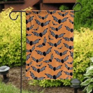May include: Orange garden flag with a repeating pattern of black bats and the words "FUCK TRUMP" in a bat-shaped design. The flag is hanging on a black metal stand in a garden setting.