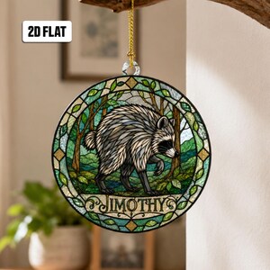 May include: A circular hanging ornament featuring a stained glass style illustration of a raccoon in a forest setting. The design includes a decorative border with green leaves and the name JIMOTHY printed in black text at the bottom center.