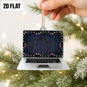 May include: A flat, laptop-shaped Christmas ornament featuring a screen displaying colorful lines of computer code framed by small, glowing holiday lights. The silver-colored laptop base includes a black trackpad with the name JACKSON printed.