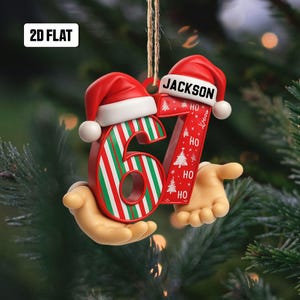 May include: A Christmas ornament shaped like the number 67, held by two hands. The number 6 is red with green and white stripes, and the number 7 is red with white Christmas trees and the word "HO". Both numbers have a red Santa hat.