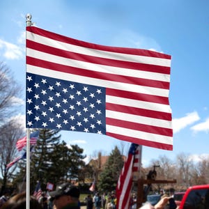 May include: A red, white, and blue American flag with the stars on the wrong side. The flag is flying on a pole in front of a crowd of people.