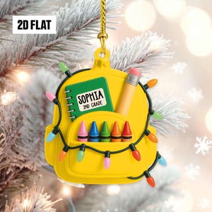 May include: A yellow backpack ornament with a pencil, crayons, and a notebook that reads "SOPHIA 2ND GRADE." The ornament is decorated with colorful Christmas lights and a gold hanging loop. The text "2D FLAT" is in the upper left corner.