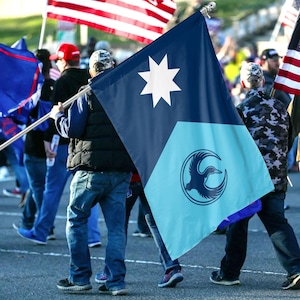 May include: A flag with a dark blue upper section featuring a white eight-pointed star and a light blue lower section with a circular emblem of a bird. The flag is being carried by a person in a crowd, with other flags visible in the background.