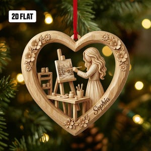 May include: A heart-shaped wooden ornament with a carved floral border and a central scene of a person painting a landscape. The name "Sophia" is inscribed on the ornament. The text "2D FLAT" is in the upper left corner. A red ribbon is attached for hanging.