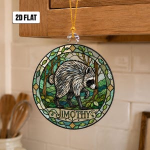 May include: A circular hanging ornament featuring a stained glass style illustration of a raccoon in a forest setting. The raccoon is grey and white with black markings. The text JIMOTHY is printed at the bottom. It hangs from a gold string.