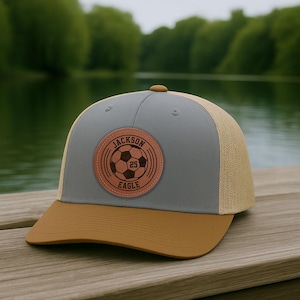 May include: A gray and tan baseball cap with a brown brim. The cap features a circular leather patch with the text "JACKSON EAGLE 25" around a soccer ball graphic. The hat is sitting on a wooden surface.