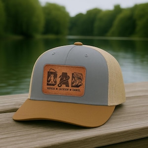 May include: A gray and tan baseball cap with a brown brim. The cap features a brown leather patch with the word "DAD" and fishing-themed graphics. The names "SOPHIA", "JACKSON", and "DANIEL" are printed below.