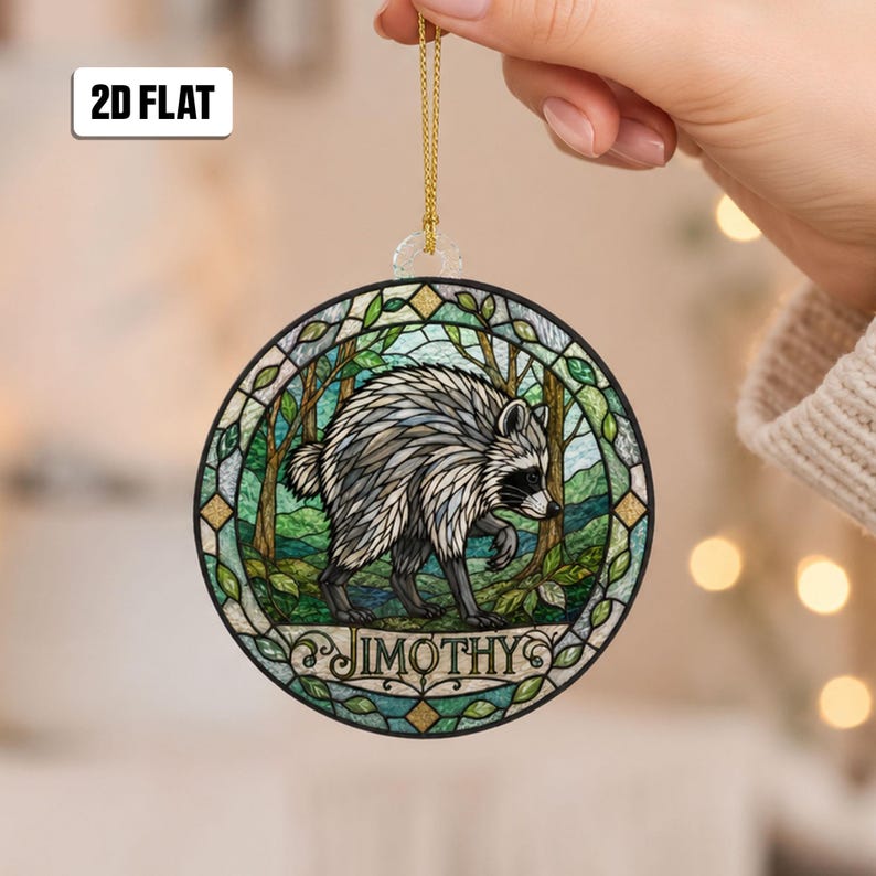 May include: A circular hanging ornament featuring a stained glass style illustration of a raccoon in a forest setting. The raccoon is grey, black, and white. The name JIMOTHY is printed in black text at the bottom. It includes a gold hanging cord.