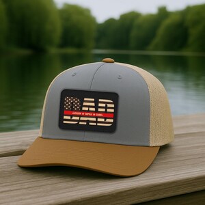 May include: A gray and tan baseball cap with a brown brim. The cap features a rectangular patch with the word "DAD" and the names "Jackson Sophia Daniel" in a patriotic design with a red stripe.