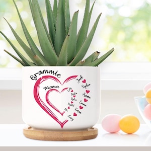 May include: White ceramic planter with a pink heart design and the words "Gramme mama" on the front. The heart is surrounded by names, including Jack, Sophia, Anna, Emma, and Lucy.