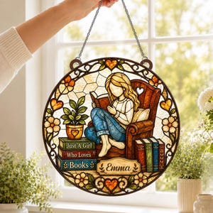 May include: A circular stained glass suncatcher featuring a person reading a book in a chair. The design includes a stack of books labeled "Just A Girl Who Loves Books" and a nameplate reading "Emma," surrounded by floral and heart motifs.