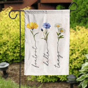 May include: A white garden flag with three different flowers and their names written underneath: foxtrot, delta, tango.