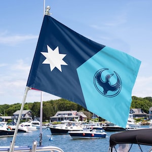 May include: A navy blue and light blue flag with a white star and a bird emblem. The flag is displayed on a boat, with other boats and houses in the background. The flag is waving in the wind.