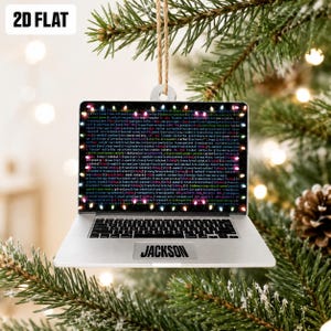 May include: A flat, silver-colored laptop ornament featuring a screen display of dense computer code framed by colorful Christmas lights. The name JACKSON is printed in black on the palm rest area. The ornament hangs from a tan twine loop.