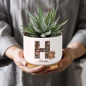 May include: White ceramic planter with a bookshelf design forming the letter H. The planter has a wooden base and is filled with a green succulent plant. The planter is personalized with the name "Hannah".