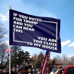 May include: A navy blue flag with white text that reads "IF YOU VOTED FOR TRUMP AND YOU CAN READ THIS YOU ARE TOO CLOSE TO MY HOUSE." The flag is mounted on a silver pole and is waving in the wind.