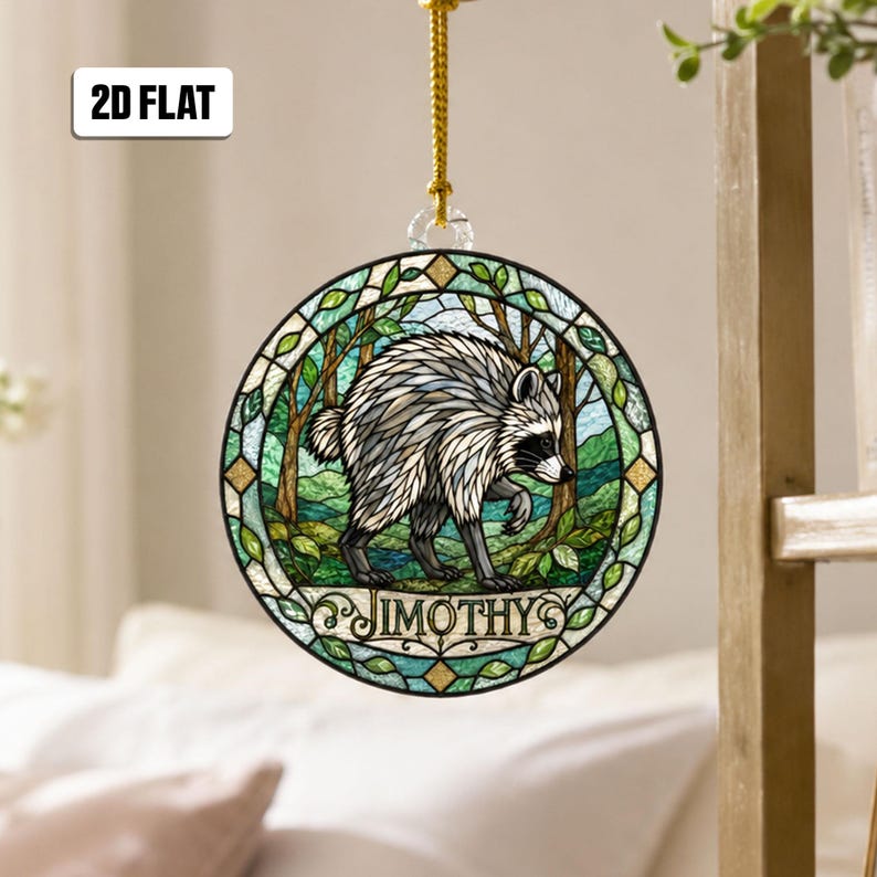 May include: A circular hanging ornament featuring a stained glass style illustration of a raccoon in a forest. The design includes green foliage, a blue sky, and the name JIMOTHY printed in black letters at the bottom. A gold cord is attached.