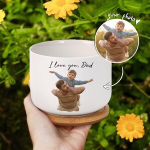 May include: White ceramic planter with a wood base. The planter has a photo of a father and son printed on it. The photo shows the father carrying the son on his back. The son is flying with his arms outstretched. The text "I love you, Dad" is printed on the planter.