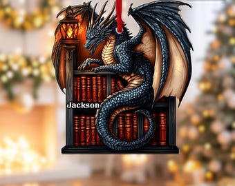 May include: A dragon-themed Christmas ornament with a red ribbon for hanging. The ornament features a detailed illustration of a dragon perched on a bookshelf, with the name "Jackson" visible. The dragon has blue scales and large wings.