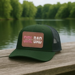 May include: A dark green trucker hat with a mesh back and a flat brim. The hat features a rectangular patch with the words "GIRL DAD" in pink and white, with the names "DANIEL SOPHIA JACKSON" below.