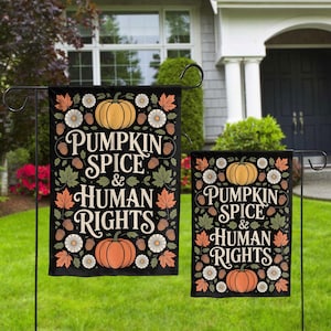 May include: Two black garden flags with the text "PUMPKIN SPICE & HUMAN RIGHTS". The flags feature orange pumpkins, white daisies, acorns, and fall leaves. The flags are displayed on black metal stands in a front yard.