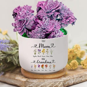 May include: White ceramic planter with the text "First Mom Now Grandma" and a floral design. The planter has a wooden base and is filled with purple succulents.