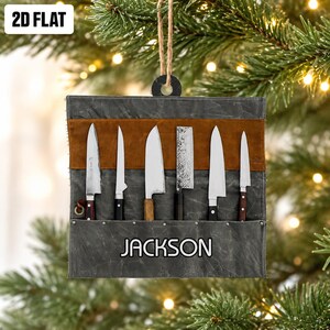 May include: A flat, square-shaped hanging ornament featuring a dark gray canvas knife roll with a brown leather accent strip. Six silver-colored metal knives are tucked into the roll. The name JACKSON is printed in white letters on the front.