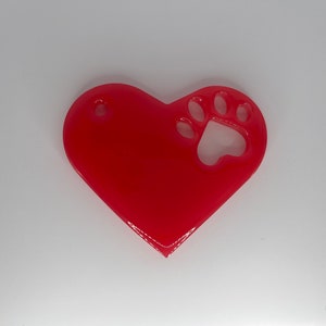 May include: A red heart-shaped charm with a paw print cut out. The charm has a hole at the top for attaching to a keychain or necklace.