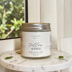 Puede incluir: Un frasco de vidrio transparente con tapa de metal plateado que contiene el acondicionador sin enjuague Living Water Organics Tallow & Shea. La etiqueta blanca presenta texto negro que dice Living Water Organics, Tallow & Shea, Leave-in Conditioner.