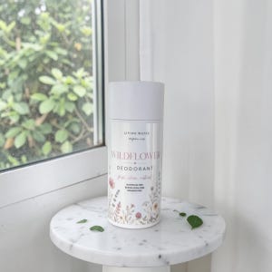 Könnte beinhalten: Ein weißer, zylindrischer Deodorant-Behälter mit Verschlusskappe, bedruckt mit Living Water Organics, Wildflower und Deodorant. Das Etikett zeigt Illustrationen kleiner bunter Blumen und den Hinweis auf Aluminium-, Natron- und Parabenfreiheit.