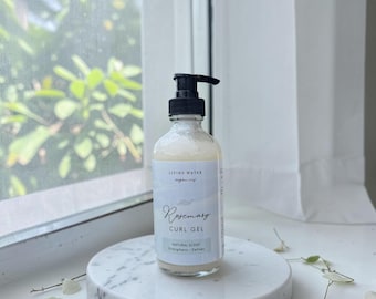 May include: A clear glass bottle of "Rosemary Curl Gel" with a black pump dispenser sits on a white marble tray. The label reads "Living Water Organics" and "Natural Scent". The background shows a window with greenery and a white curtain.