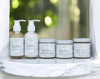Op de afbeelding: Een collectie van vijf haarverzorgingsproducten, waaronder twee pompflessen met het label Tallow Shampoo en Tallow Conditioner, en drie potten met het label Tallow Deep Conditioner, Tallow Curl Cream en Tallow Honey Curl Gel, allemaal voorzien van witte etiketten.