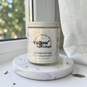 Puede incluir: Un tarro de cristal de Tallow Curl Cream con tapa plateada sobre una bandeja de mármol blanco. La etiqueta dice "Living Water Organics Tallow Curl Cream" y "Hydrating Curl Cream". El tarro está etiquetado "Peso neto 226 g".