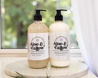Può includere: Due flaconi di prodotti per la cura dei capelli con dispenser a pompa neri. Uno contiene un liquido ambrato chiaro, l'altro un liquido bianco. Entrambi sono etichettati "Aloe & Tallow" e contengono 473 ml. Le etichette recitano anche "Shampoo Idratante" e "Leave-In Conditioner."