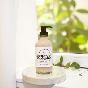 Puede incluir: Una botella de vidrio transparente con un dispensador de bomba negro, etiquetada como "Rosemary & Flaxseed Gel". La botella contiene un líquido beige claro y está sobre una superficie de mármol blanco. La etiqueta menciona semillas de lino y agua de romero.