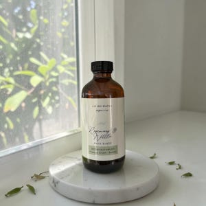 Könnte beinhalten: Eine braune Glasflasche mit Rosemary & Nettle Hair Rinse und schwarzem Verschluss auf einem weißen Marmortablett. Das Etikett lautet "Living Waters Organics" und "Botanically Infused". Die Flasche steht in der Nähe eines Fensters mit grünem Laub.
